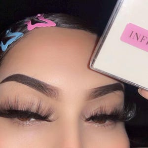 Puede incluir: Primer plano de ojos con pestañas largas y oscuras y cejas definidas. Se ven pinzas para el pelo con forma de mariposa. Una caja con la palabra "INFINITE" en una etiqueta rosa se sostiene en el encuadre.