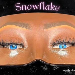 May include: A close-up of a brown face mask with blue eyes and long black eyelashes. The word "Snowflake" is written in pink cursive above the eyes. The website address angelhustle.etsy.com is written in white at the bottom.