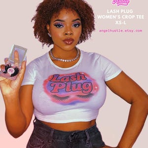 Pode incluir: Um top curto branco com o texto "Lash Plug" em um design rosa e roxo. A imagem também mostra uma mulher segurando um estojo de cílios postiços. O texto "Angel Hustle" e o endereço do site também são visíveis.