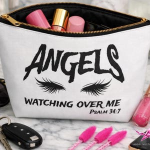 Könnte beinhalten: Weiße Kosmetiktasche mit schwarzem Reißverschluss und dem Wort "ANGELS" in fetten Buchstaben. Darunter schwarze Wimpern und der Text "WATCHING OVER ME" und "PSALM 34:7". Die Tasche ist mit Make-up gefüllt.