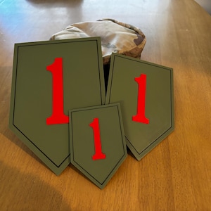May include: Three olive green shield-shaped plaques with a red number 1. The plaques are of varying sizes and have a black border. A camouflage hat rests behind the plaques on a wooden surface.