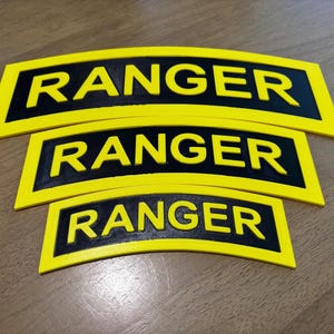 May include: Three yellow and black Ranger patches are stacked on a wooden surface. Each patch has a black rectangular base with the word "RANGER" in bold yellow letters. The patches are slightly curved.