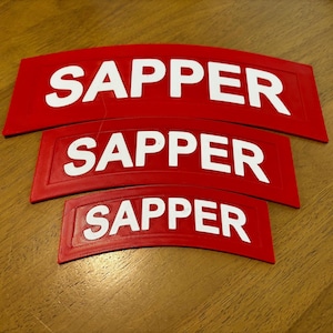 May include: Three red patches with the word "SAPPER" in white block letters. The patches are curved and vary in size, with the largest at the top and the smallest at the bottom. The patches are on a wooden surface.