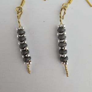 Könnte beinhalten: Ein Paar handgefertigte Ohrringe mit goldfarbenen Haken und Draht. Jeder Ohrring hat einen vertikalen Strang aus dunkelgrauen Perlen, die durch kleine silberne Perlen getrennt sind. Die Ohrringe sind ein schlichtes, elegantes Design.