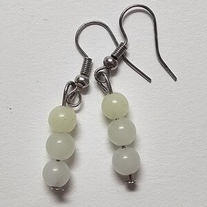 May include: A pair of dangle earrings featuring three pale green, round beads suspended from silver-tone hooks. The beads have a subtle, pearlescent sheen. The earrings are simple and elegant, suitable for everyday wear.