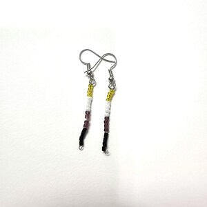 Puede incluir: Un par de pendientes colgantes con ganchos plateados. Cada pendiente presenta una hebra vertical de pequeñas cuentas de colores. Las cuentas están dispuestas en secciones amarillas, blancas, moradas y negras, creando un patrón de rayas.