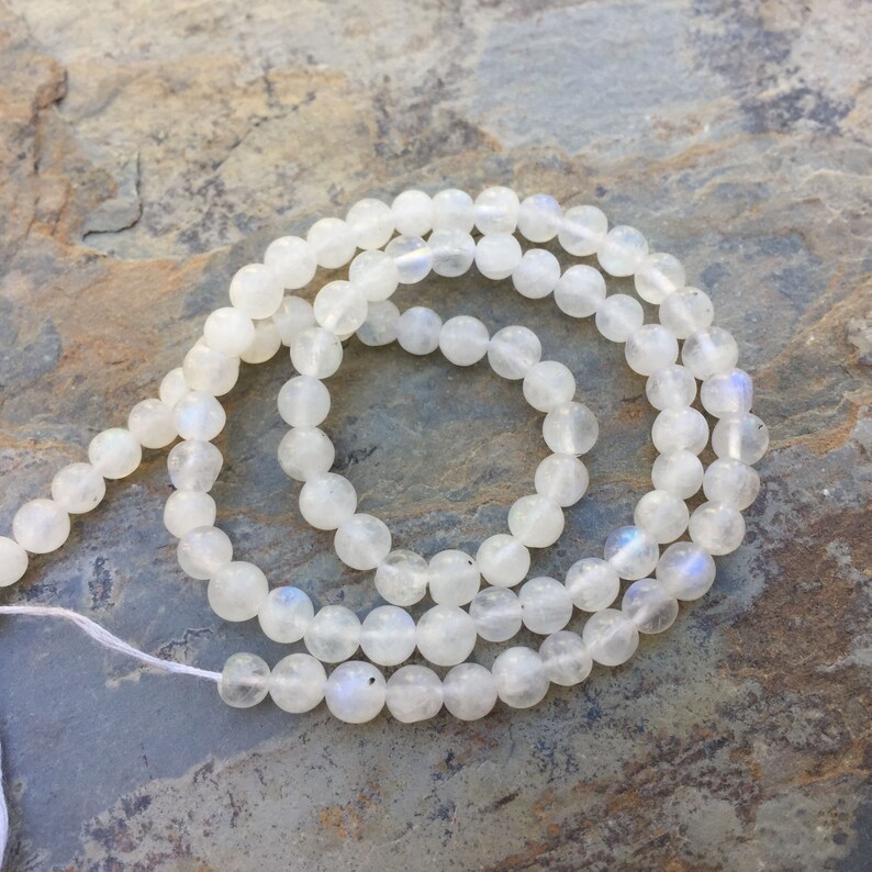 May include: A strand of white moonstone beads, approximately 8mm in diameter, with a subtle iridescent sheen. The beads are strung on a white thread and are arranged in a loose circle.