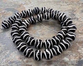 May include: A strand of spherical beads featuring a glossy black finish with a continuous, winding white stripe pattern. The beads are arranged in a circular formation, strung together on a thin, light-coloured cord, showcasing a uniform design.
