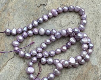 May include: A strand of lavender-colored freshwater pearls. The pearls are round and have a slight iridescent sheen. The pearls are strung on a thin, purple thread.
