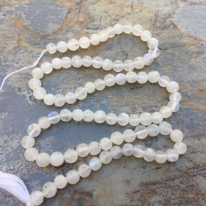 May include: A strand of white moonstone beads, each bead is round and smooth. The beads are strung on a white thread and have a white tassel at one end.