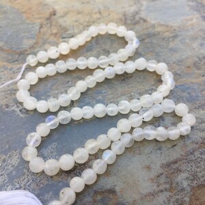 May include: A strand of white moonstone beads. The beads are round and have a smooth, polished finish. The beads are strung on a white thread.