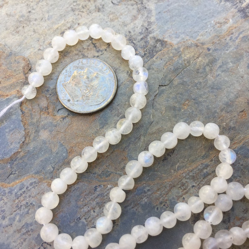 May include: A strand of white moonstone beads, approximately 6mm in diameter, displayed on a textured grey surface. A UK 20 pence coin is included for size reference.