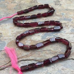 May include: A strand of dark red gemstone beads, each bead is rectangular and faceted. The beads are strung on a cord with a pink tassel at one end.