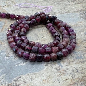 May include: Strands of faceted, deep burgundy gemstone beads. The beads are a mix of dark red and purple hues, strung together with a thin, light purple cord. The beads have a geometric, multi-faceted shape, reflecting light.