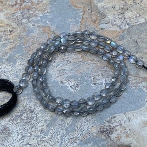 May include: A strand of gray faceted labradorite beads with a black tassel. The beads are oval shaped and have a slight iridescent sheen.