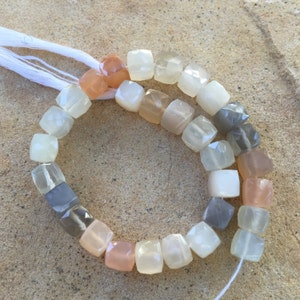 May include: A strand of faceted gemstone beads in shades of white, gray, and peach. The beads are square-shaped and strung on a white thread.