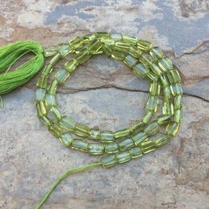 Peut inclure: Un brin de perles de péridot vert clair, chaque perle est de forme rectangulaire. Les perles sont enfilées sur un fil vert avec un pompon vert à une extrémité.