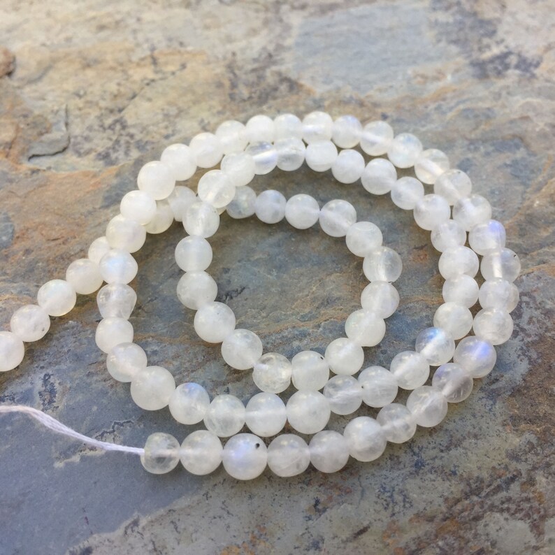 May include: A strand of white moonstone beads, each bead is round and smooth. The beads are arranged in a circular pattern on a textured surface.