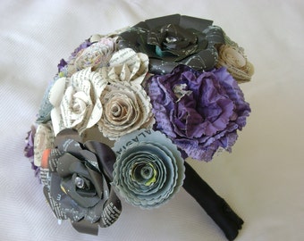 May include: A bouquet of paper flowers made from book pages and fabric. The bouquet features a variety of colors, including purple, blue, green, and white. The flowers are arranged in a circular shape and are held together by a black ribbon.