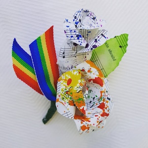 May include: A rainbow ribbon attached to two paper roses made from sheet music with colorful paint splatters. The roses are arranged in a bouquet with a green leaf. The leaf has the text "ain should" written on it.
