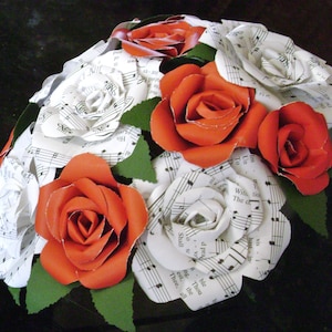 May include: A bouquet of paper flowers made from sheet music. The flowers are red and white and are arranged in a circular shape. The bouquet is surrounded by green leaves.
