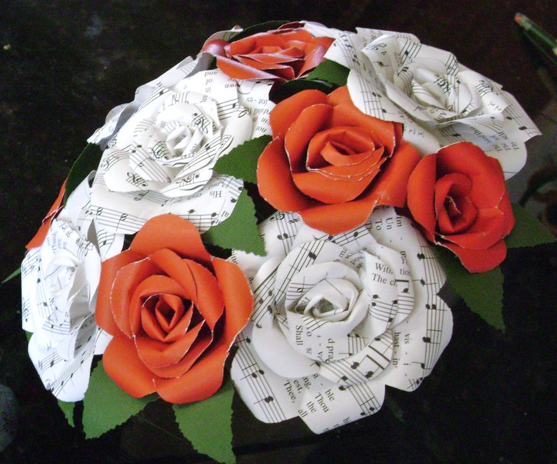 May include: A bouquet of paper flowers made from sheet music. The flowers are red and white and are arranged in a circular shape. The bouquet is surrounded by green leaves.