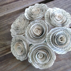 May include: A bouquet of paper flowers made from book pages. The flowers are white and have a delicate, layered appearance. The flowers are arranged in a cluster and have a vintage aesthetic.