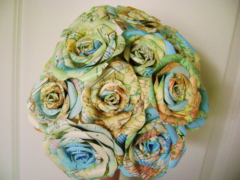 May include: A bouquet of paper roses made from maps in shades of blue, green, and yellow. The roses are arranged in a circular shape and are tied together with a ribbon.