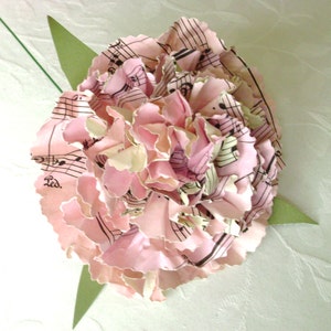 Peut inclure: Une fleur en papier rose réalisée à partir de partitions musicales. La fleur est composée de nombreuses couches de papier, et les notes de musique sont visibles sur les pétales. La fleur est sur un fond blanc.