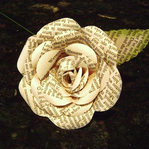 May include: A white paper rose made from pages of a book. The rose is made of several layers of paper, each with text printed on it. The rose is sitting on a dark brown surface.