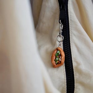 May include: A miniature, handmade food charm in the shape of a baked pastry, attached to a silver-colored keychain. The charm is brown with green and white details, hanging from a black zipper on a cream-colored jacket.
