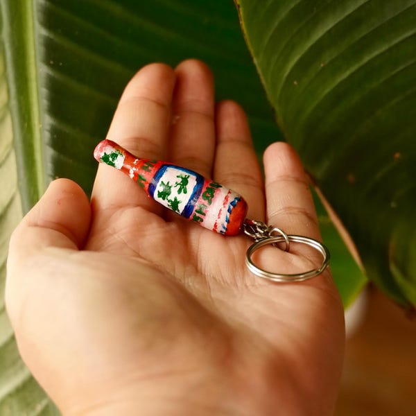 May include: A miniature, hand-painted bottle-shaped keychain. The keychain features a red, white, and blue design with green shamrocks. The keychain is attached to a silver-colored keyring. The background includes a large green leaf.