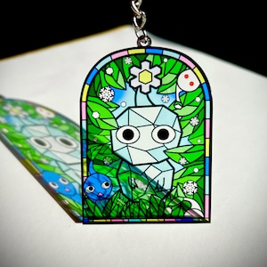 Peut inclure: Un porte-clés de style vitrail représentant un personnage géométrique aux grands yeux, entouré de feuillage vert, d'une créature bleue et d'une fleur blanche. Le design est encadré d'une bordure noire et d'une chaîne argentée.