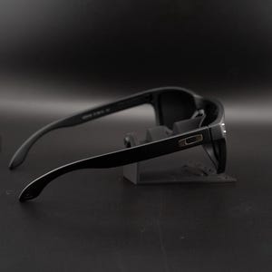 May include: Black sunglasses with a rectangular frame and curved temples. The sunglasses are displayed on a gray stand against a dark background. The lenses are dark, and the frame has a matte finish.