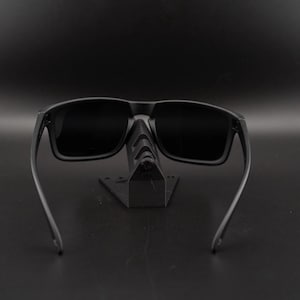 May include: Black sunglasses with a rectangular frame and dark lenses. The sunglasses are displayed on a black stand against a black background. The arms of the sunglasses extend outward.
