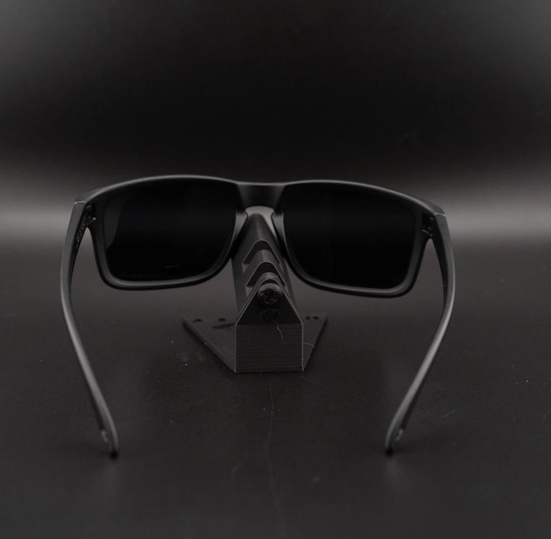 May include: Black sunglasses with a rectangular frame and dark lenses. The sunglasses are displayed on a black stand against a black background. The arms of the sunglasses extend outward.