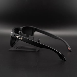 May include: Black sunglasses with a matte finish. The sunglasses have a rectangular frame and dark lenses. The temples are sleek and curve down towards the ear. A small silver accent is visible on the side of the frame.
