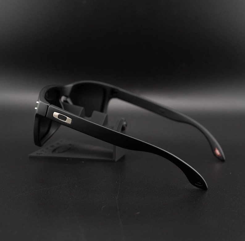 May include: Black sunglasses with a matte finish. The sunglasses have a rectangular frame and dark lenses. The temples are sleek and curve down towards the ear. A small silver accent is visible on the side of the frame.