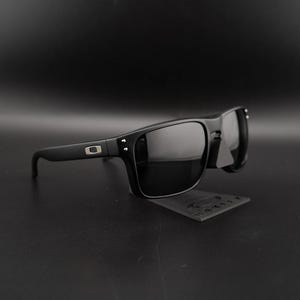 May include: Black sunglasses with a rectangular frame and dark lenses. The sunglasses have a matte finish and a small silver logo on the temple. The sunglasses are displayed on a black surface.