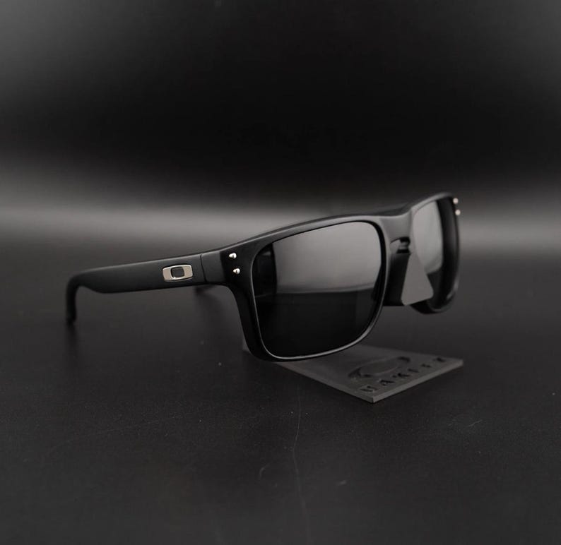 May include: Black sunglasses with a rectangular frame and dark lenses. The sunglasses have a matte finish and a small silver logo on the temple. The sunglasses are displayed on a black surface.