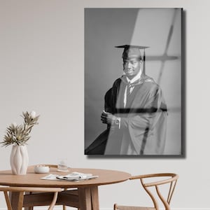 Peut inclure: Photographie en noir et blanc d'une personne en tenue de remise de dipl&ocirc;me, comprenant une coiffe, une toge et une cravate. La personne sourit et pose, les bras crois&eacute;s. L'arri&egrave;re-plan est de couleur grise unie.
