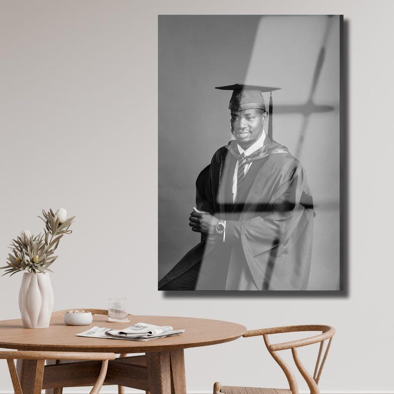 Peut inclure: Photographie en noir et blanc d'une personne en tenue de remise de dipl&ocirc;me, comprenant une coiffe, une toge et une cravate. La personne sourit et pose, les bras crois&eacute;s. L'arri&egrave;re-plan est de couleur grise unie.