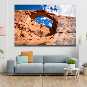 Peut inclure: Une grande impression encadr&eacute;e d'une arche rocheuse naturelle sur fond de ciel bleu avec des nuages blancs. L'arche est de couleur brun rouge&acirc;tre. L'impression est affich&eacute;e au-dessus d'un canap&eacute; gris avec des coussins color&eacute;s.