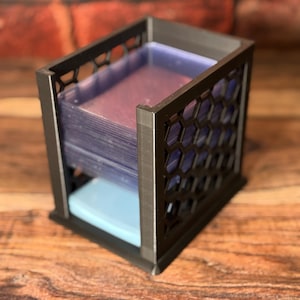 May include: A black, geometric card holder with a honeycomb design on the sides. The holder contains a stack of clear card sleeves and a light blue base. The holder is made of a dark material and sits on a wooden surface.
