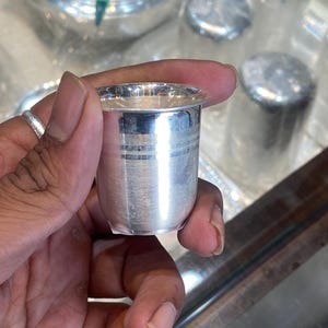 Puede incluir: Un pequeño vaso cilíndrico de plata con acabado pulido y un sutil detalle de banda horizontal cerca del borde. El recipiente presenta una superficie metálica lisa y un borde superior ligeramente ensanchado que refleja la luz.
