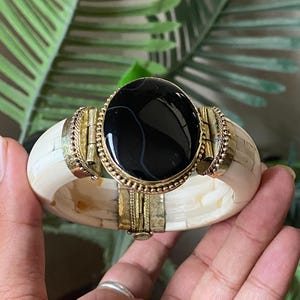 Puede incluir: Una pulsera de brazalete con bisagras que presenta una gran piedra central ovalada de color negro pulido con vetas blancas sutiles. La banda está construida con segmentos lisos de color blanco roto unidos por bisagras y monturas de metal dorado.