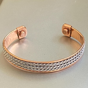 May include: A copper cuff bracelet featuring a polished copper band with three parallel silver-toned twisted wire strands running through the center. The bracelet has an open design with two circular magnetic components attached to the inner ends.