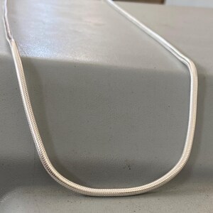 Puede incluir: Un collar de cadena de serpiente en tono plateado con un diseño liso, flexible y redondeado con un acabado metálico pulido. La cadena está dispuesta en forma de U, mostrando sus eslabones uniformes y estrechamente tejidos.