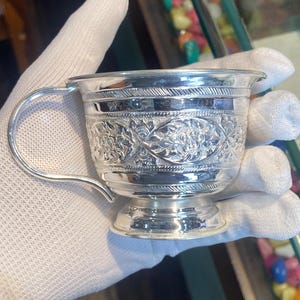 Puede incluir: Una taza de plata pulida con un intrincado patrón floral en relieve en el exterior, un asa curva y un borde acampanado. La superficie metálica es altamente reflectante y muestra una artesanía detallada con bandas decorativas.