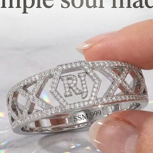 Puede incluir: Un anillo de tono plateado con un marco hexagonal central con las letras RJ, rodeado por un patrón de celosía incrustado con pequeñas piedras claras. El texto simple soul made aparece arriba y SSM.999 está grabado en la banda.